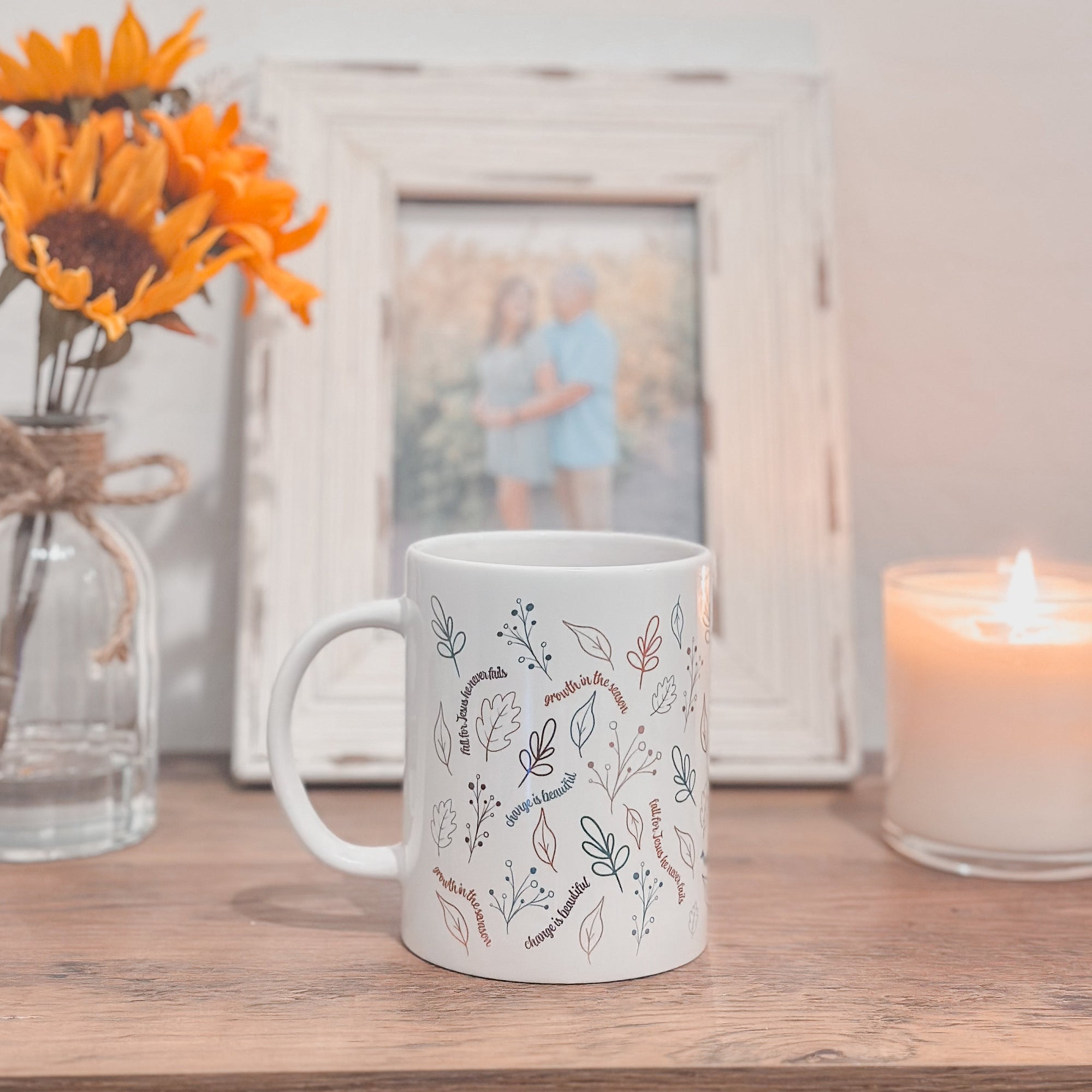 White mug with floral patterns on a wooden surface with a candle and sunflower arrangement.