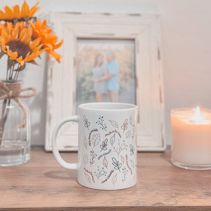 White mug with floral patterns on a wooden surface with a candle and sunflower arrangement.